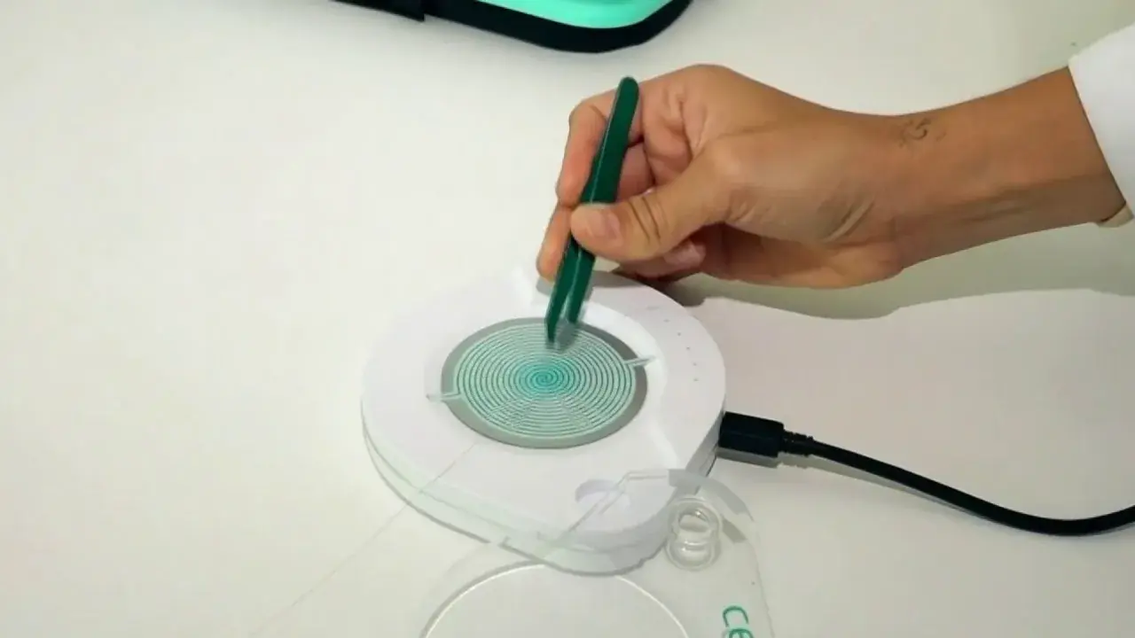Mano con pinzas verdes manipulando un dispositivo circular con espiral azul. El dispositivo está conectado a un cable negro. Las opiniones sobre el test epigenético varían.