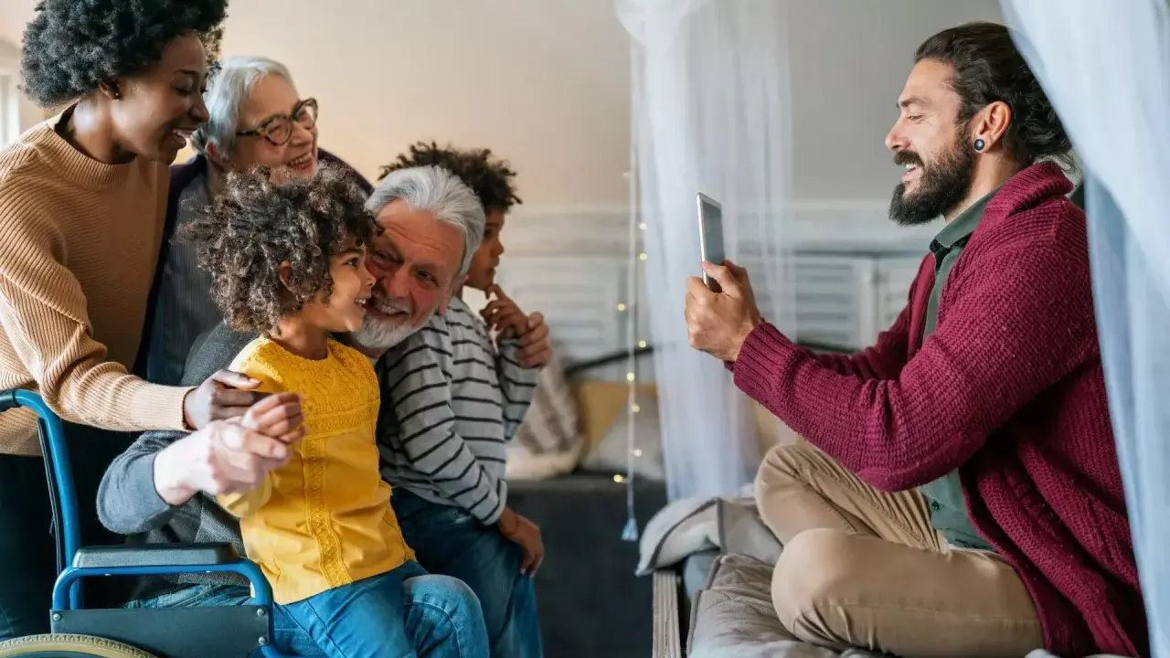 Familia multigeneracional reunida, una abuela en silla de ruedas, sonríen mientras un hombre toma una foto. Se hereda el Parkinson, pero el amor familiar perdura.