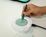 Mano con pinzas verdes manipulando un dispositivo circular con espiral azul. El dispositivo está conectado a un cable negro. Las opiniones sobre el test epigenético varían.