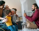 Familia multigeneracional reunida, una abuela en silla de ruedas, sonríen mientras un hombre toma una foto. Se hereda el Parkinson, pero el amor familiar perdura.