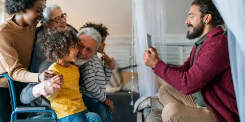 Familia multigeneracional reunida, una abuela en silla de ruedas, sonríen mientras un hombre toma una foto. Se hereda el Parkinson, pero el amor familiar perdura.