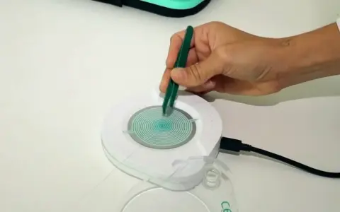 Mano con pinzas verdes manipulando un dispositivo circular con espiral azul. El dispositivo está conectado a un cable negro. Las opiniones sobre el test epigenético varían.