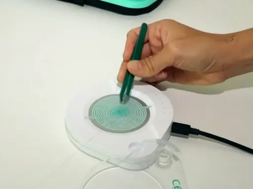 Mano con pinzas verdes manipulando un dispositivo circular con espiral azul. El dispositivo está conectado a un cable negro. Las opiniones sobre el test epigenético varían.