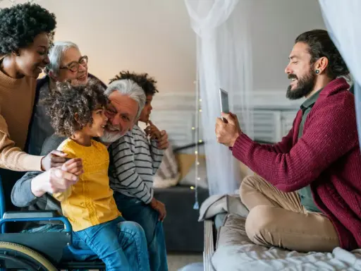 Familia multigeneracional reunida, una abuela en silla de ruedas, sonríen mientras un hombre toma una foto. Se hereda el Parkinson, pero el amor familiar perdura.