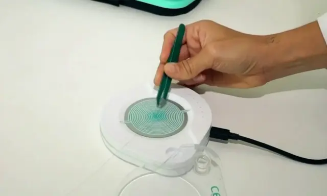 Mano con pinzas verdes manipulando un dispositivo circular con espiral azul. El dispositivo está conectado a un cable negro. Las opiniones sobre el test epigenético varían.
