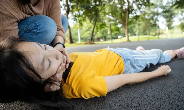 Niña inconsciente en el suelo, con una persona arrodillada a su lado, preocupada. Podría ser un síntoma de la enfermedad de Lafora.
