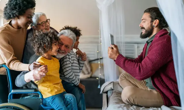 Familia multigeneracional reunida, una abuela en silla de ruedas, sonríen mientras un hombre toma una foto. Se hereda el Parkinson, pero el amor familiar perdura.
