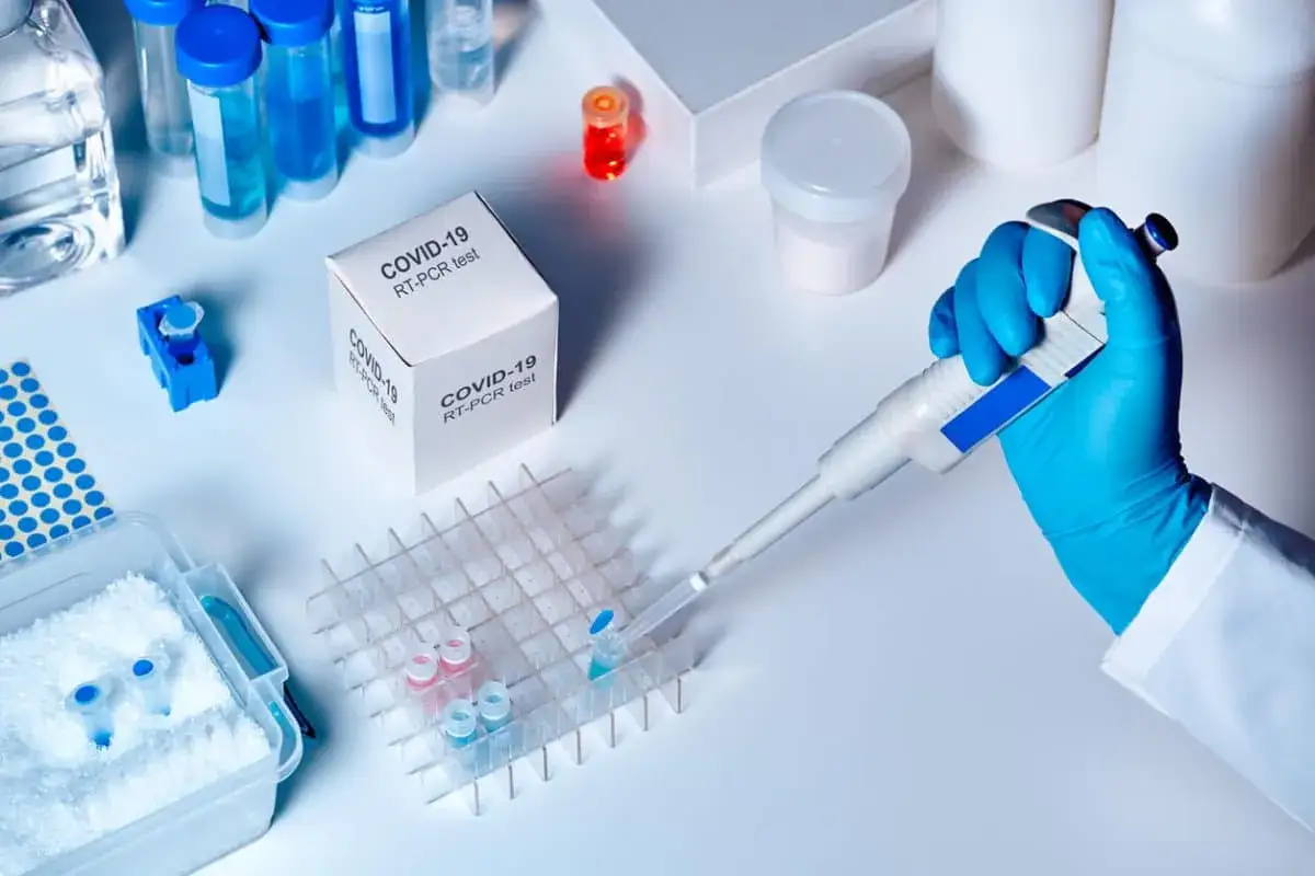 Mano enguantada usa pipeta para añadir líquido a tubos de ensayo en un laboratorio, cerca de una caja de prueba PCR COVID-19.