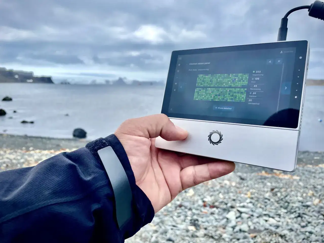Mano sostiene un dispositivo MinION Nanopore en la playa, mostrando datos de secuenciación.