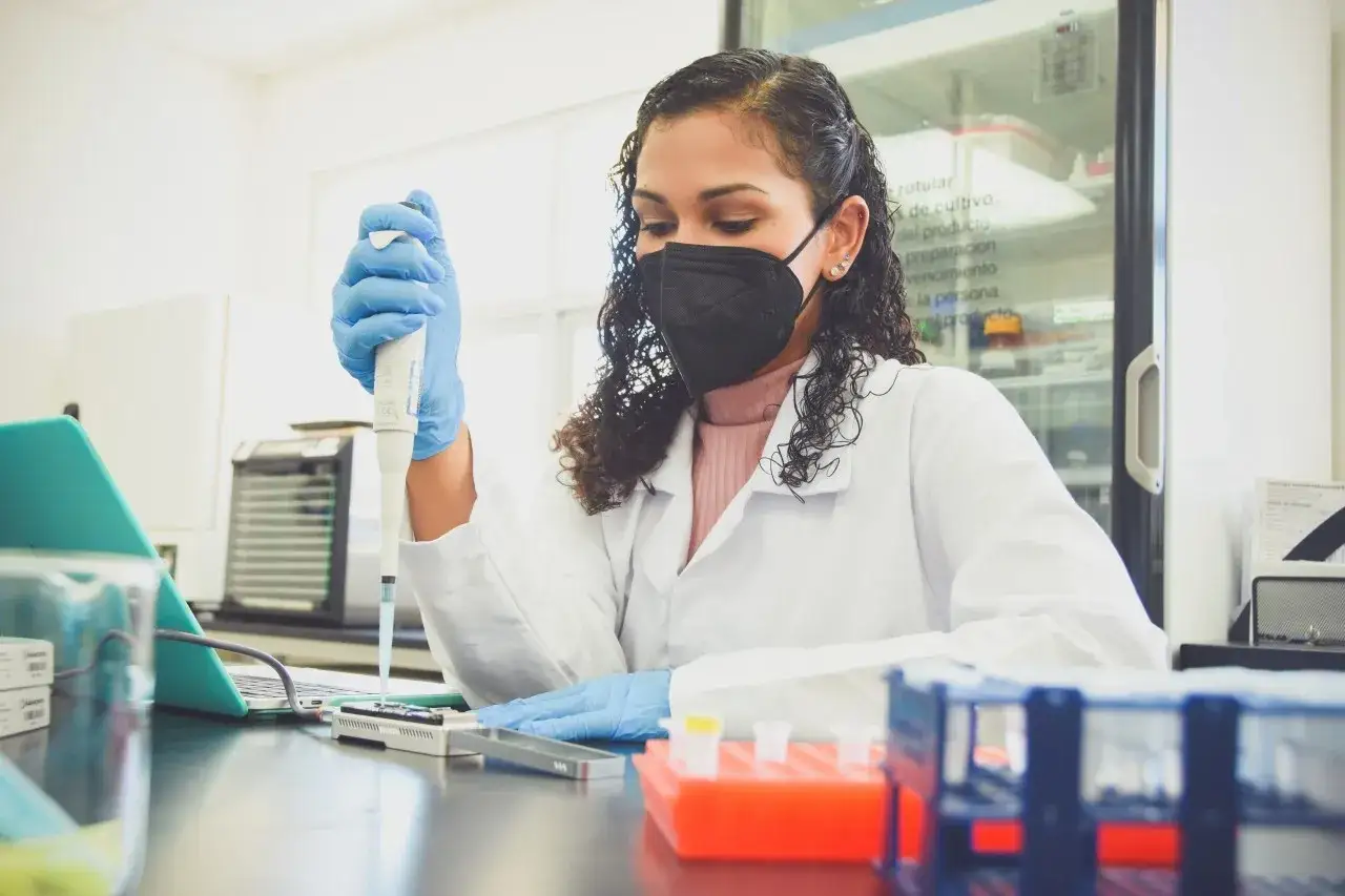 Cient&iacute;fica con mascarilla y guantes azules realiza pipeteo para secuenciaci&oacute;n ngs. Muestra tubos de ensayo y laptop en laboratorio.